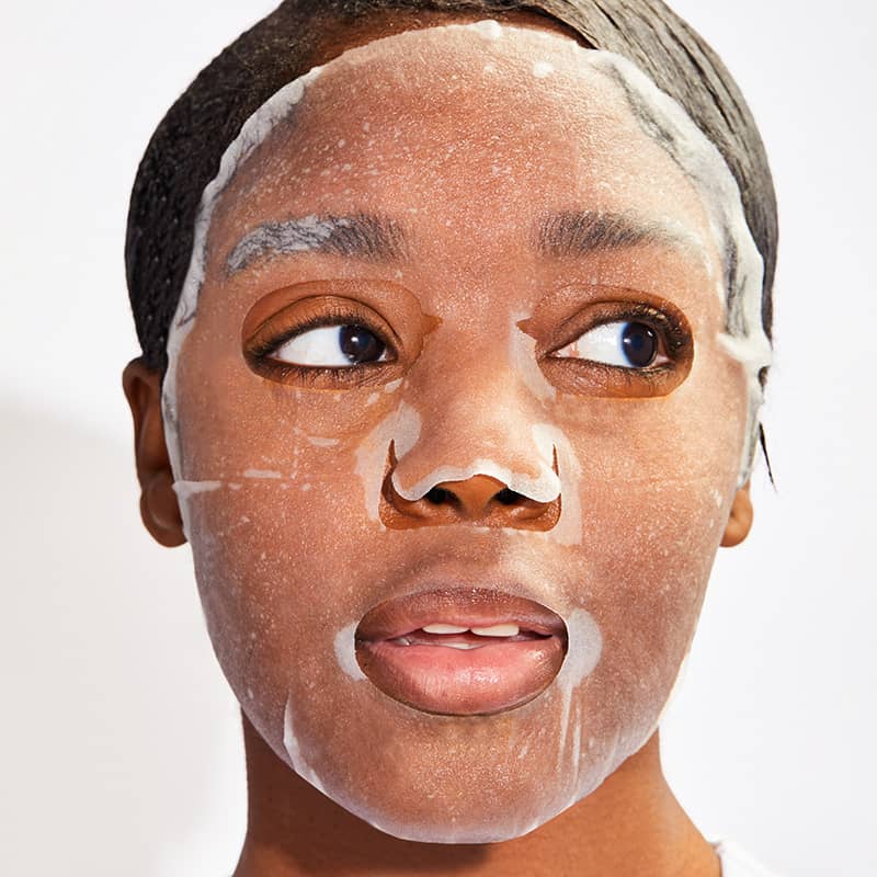 Studio photograph of a Black woman in a white studio wearing a translucent wet-textured sheet mask that highlights her side-glancing dark eyes and a subtle gap in her teeth