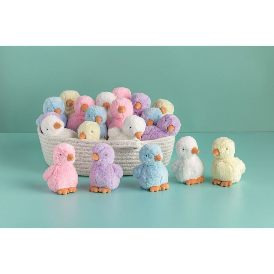 Photograph of various pastel plush ducklings, some nestled in a white woven basket and others standing individually on a teal surface, featuring soft shades of pink, purple, blue, yellow, and white with distinctive orange beaks and feet