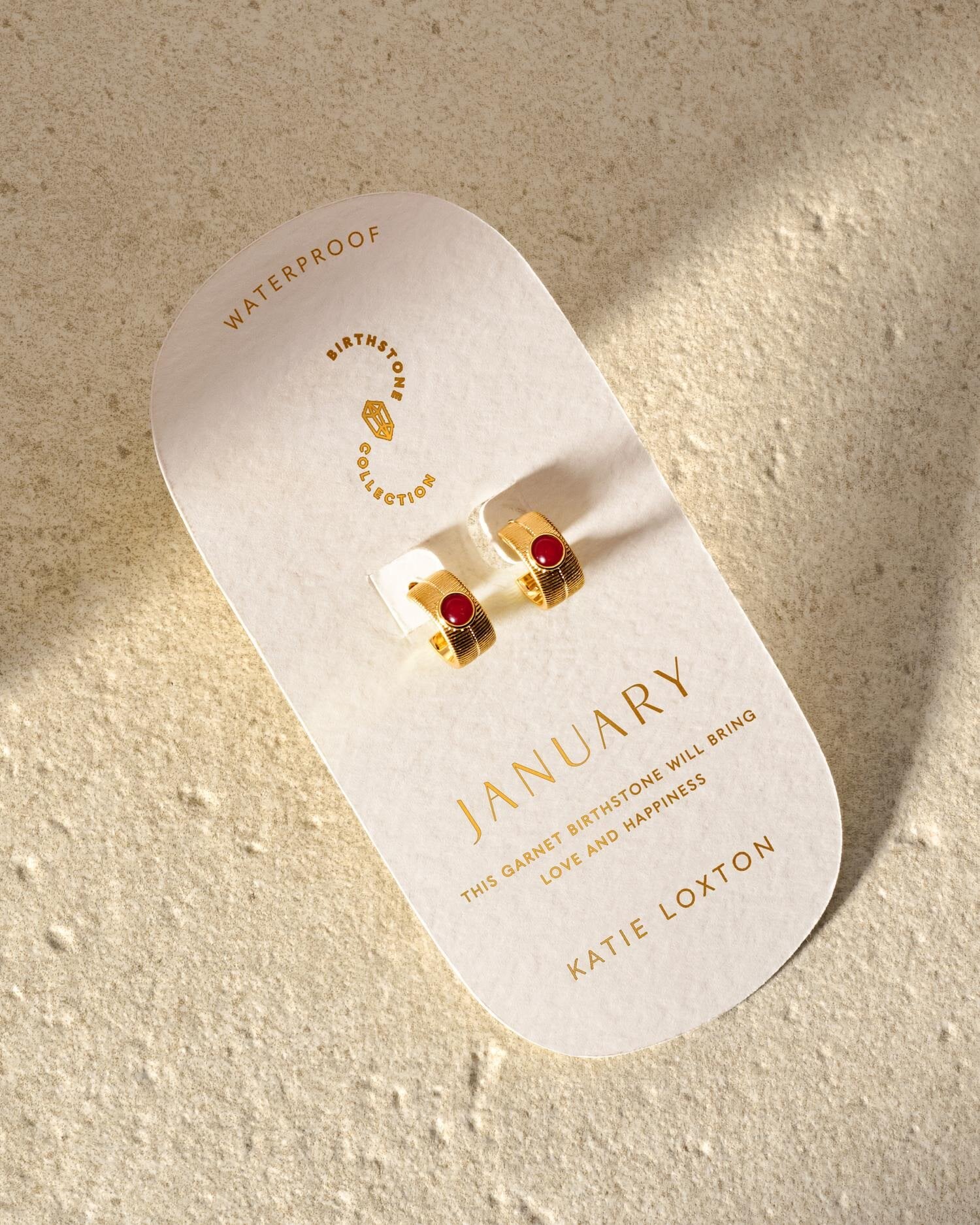 Gold hoop earrings with red garnet stones on a January birthstone display card