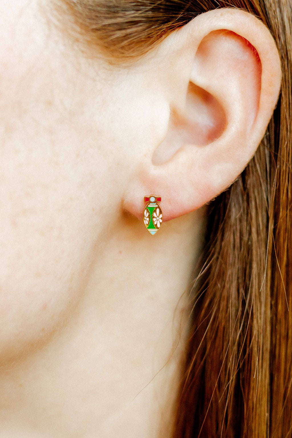 Photograph of a person's earlobe wearing a gold enamel tear-drop shaped earring featuring a vibrant green center with white floral patterns and a small red bow detail