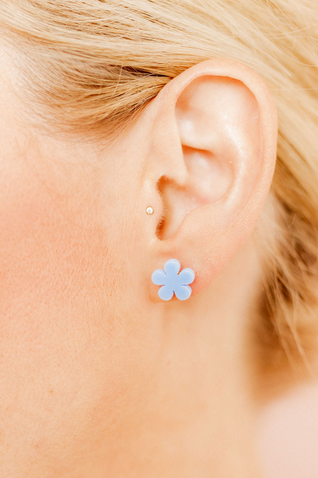 Photograph of a person's ear with blonde hair, featuring a light blue flower stud earring on the lobe and a small gold tragus piercing
