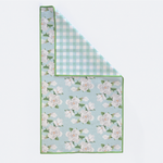 Textile kitchen towel with a white magnolia floral pattern on a light blue background, displayed on a white studio surface, featuring a lime green border and a light blue gingham pattern on the folded reverse side