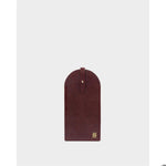 Leather eyeglass case featuring a burgundy arched pouch centered on a studio gray background with a gold-tone stud fastener on a central strap and a small gold monogram logo at the bottom corner