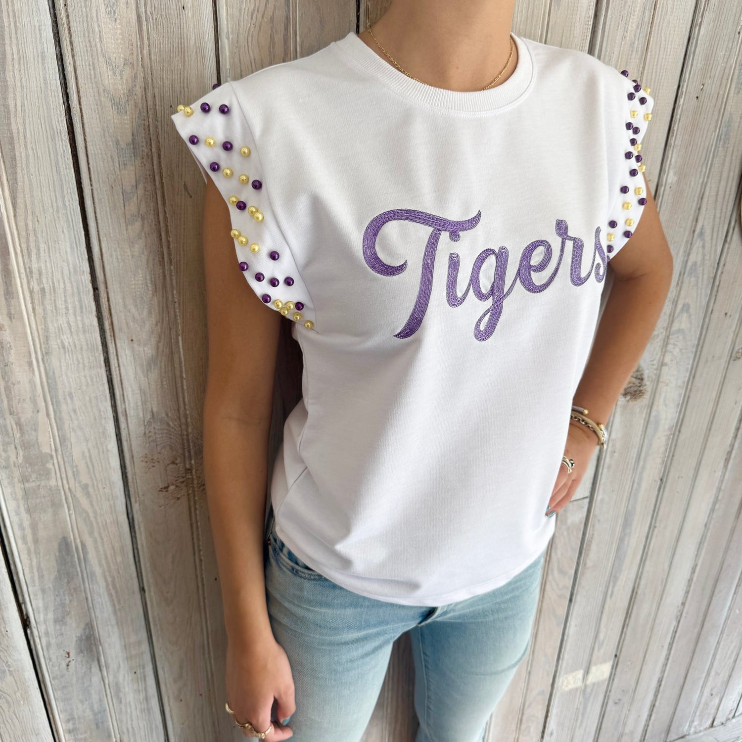 A person wears a white "Tigers" t-shirt with purple script embroidery and cap sleeves adorned with purple and gold beads, paired with light blue jeans, standing against a rustic wooden wall