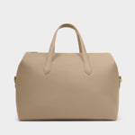 Product photograph by Katie Loxton of a taupe vegan leather weekend bag on a plain grey background featuring a gold-stamped brand logo and matching top handles with metallic hardware accents