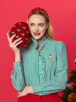 Studio fashion photograph of a blonde woman posing against a solid red background wearing a green and pink striped ruffled shirt featuring a nutcracker embroidery while holding a red velvet clutch and wearing a matching headband both adorned with tiny green Christmas trees