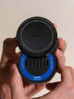 Photograph of a Pura x Capri Blue smart scent diffuser held in a person's hands, highlighting its matte black design, iconic blue Capri Blue label, and vertical scent vents