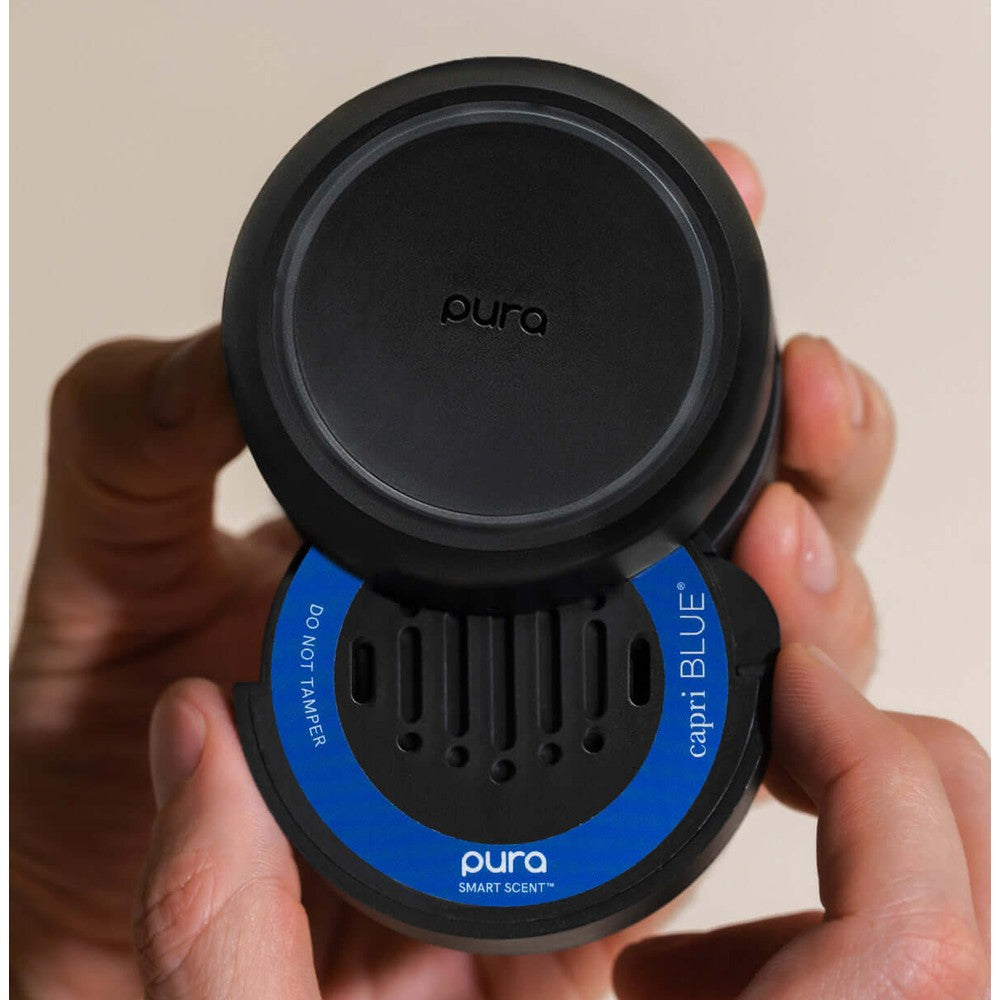 Photograph of a Pura x Capri Blue smart scent diffuser held in a person's hands, highlighting its matte black design, iconic blue Capri Blue label, and vertical scent vents