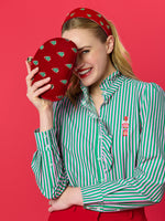 Studio photograph of a smiling blonde woman against a red background wearing a green and pink striped ruffled shirt with a pink nutcracker embroidery while holding a red velvet clutch decorated with small green Christmas trees