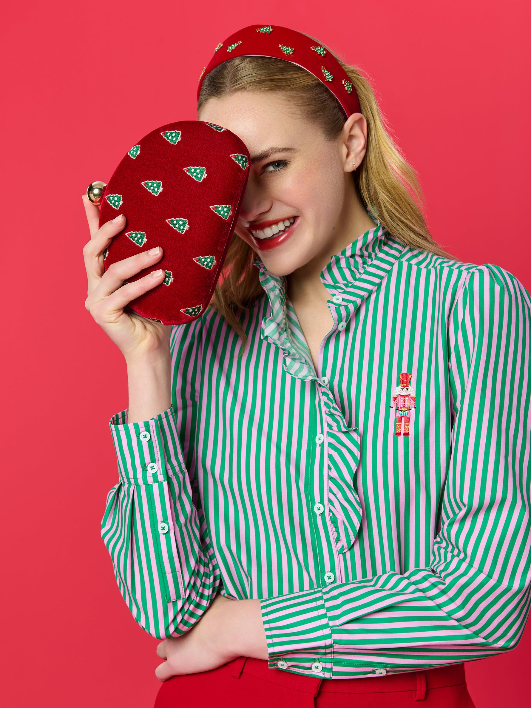 Studio photograph of a smiling blonde woman against a red background wearing a green and pink striped ruffled shirt with a pink nutcracker embroidery while holding a red velvet clutch decorated with small green Christmas trees