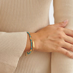 Two gold bracelets with turquoise beads and a rectangular charm worn on a wrist