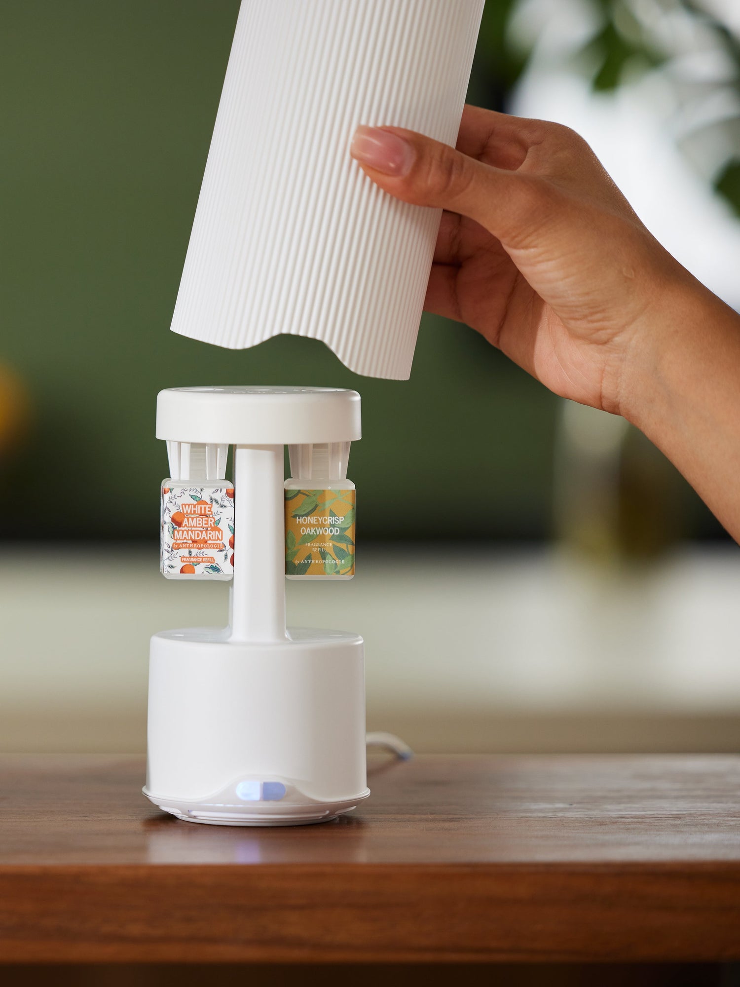 Photograph by Anthropologie of a smart home fragrance diffuser on a wooden table with a hand lifting a white fluted cover to reveal White Amber Mandarin and Honeycrisp Oakwood scent refills