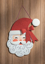 Decorative die-cut metal wall hanging of a smiling Santa Claus face on a wood-paneled background featuring an iconic red glittery hat with a large matching bow and a curly white beard with silver swirls