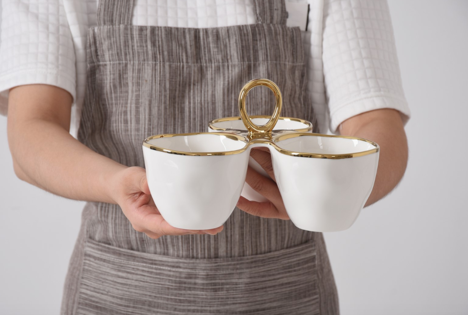 White porcelain three-compartment serving dish with gold trim and a central gold ring handle held by a person wearing a striped apron and white quilted shirt