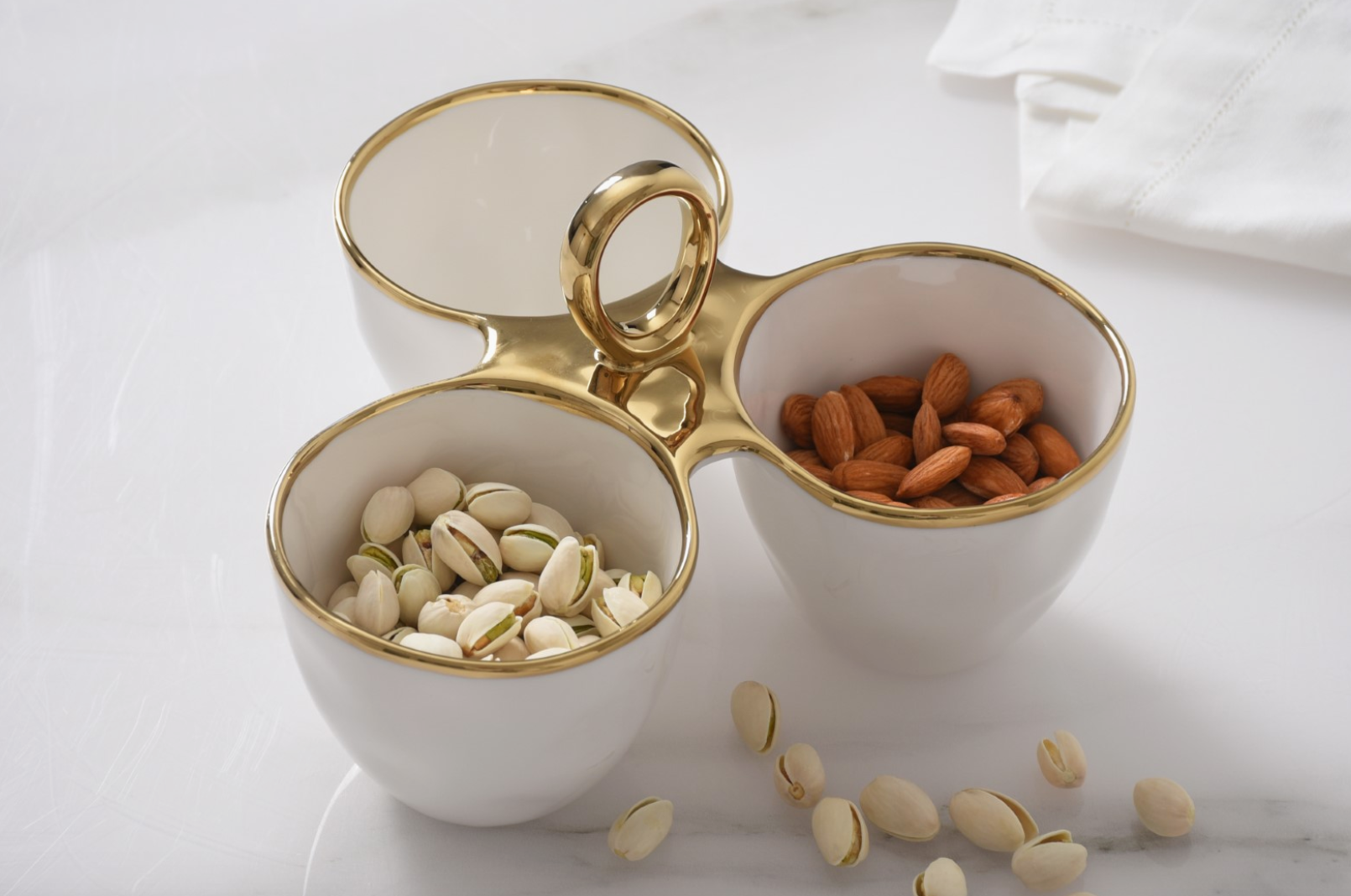 White ceramic three-section serving dish with lustrous gold trim and a central handle holds vibrant green pistachios and rich brown almonds displayed on a pristine white marble tabletop alongside scattered pistachios and a crisp white linen napkin