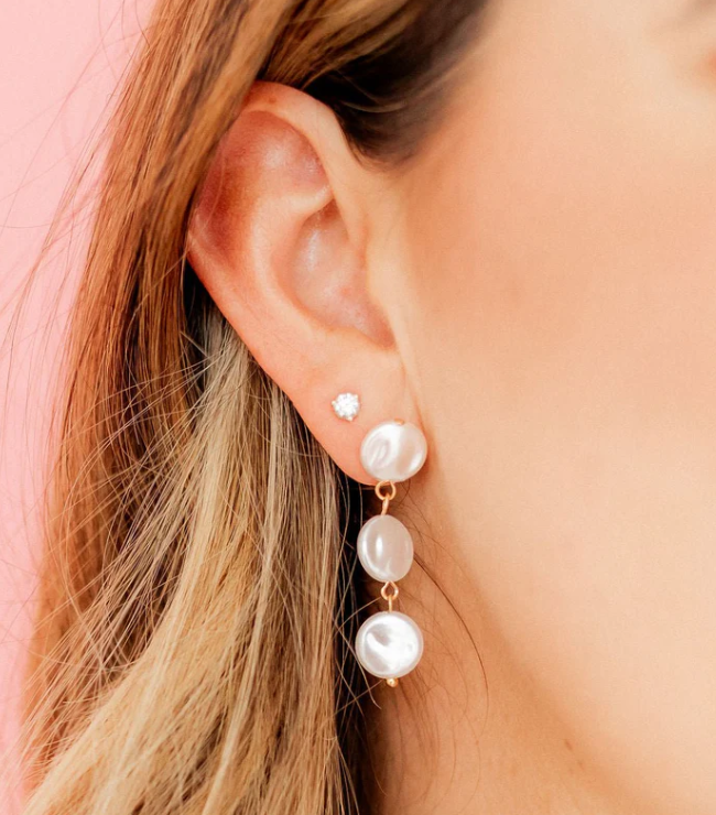Close-up of an ear adorned with a small sparkling stud earring and a longer dangle earring featuring three irregular white pearls linked by gold-toned wire set against a vibrant pink background