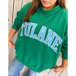 Person wearing a green 'TULANE' sweatshirt on a wooden wall.