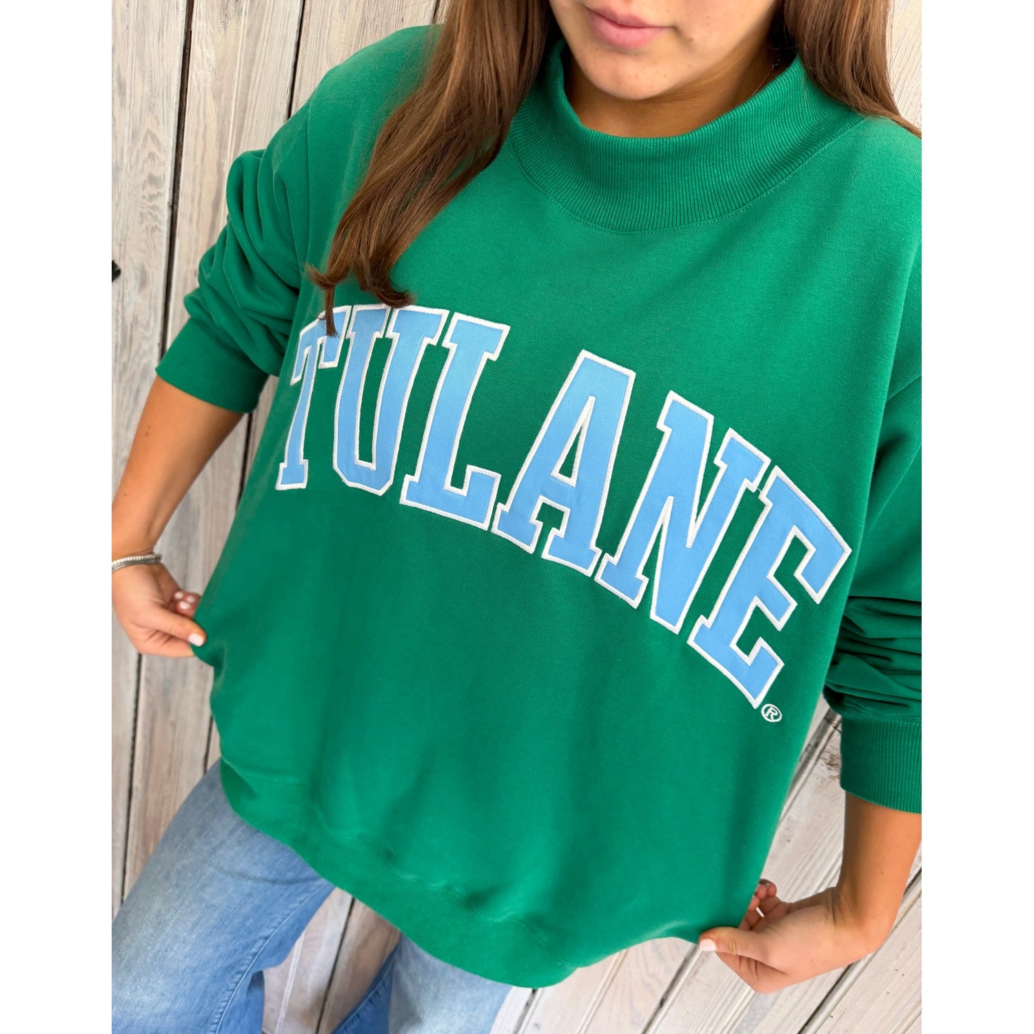 Person wearing a green 'TULANE' sweatshirt on a wooden wall.