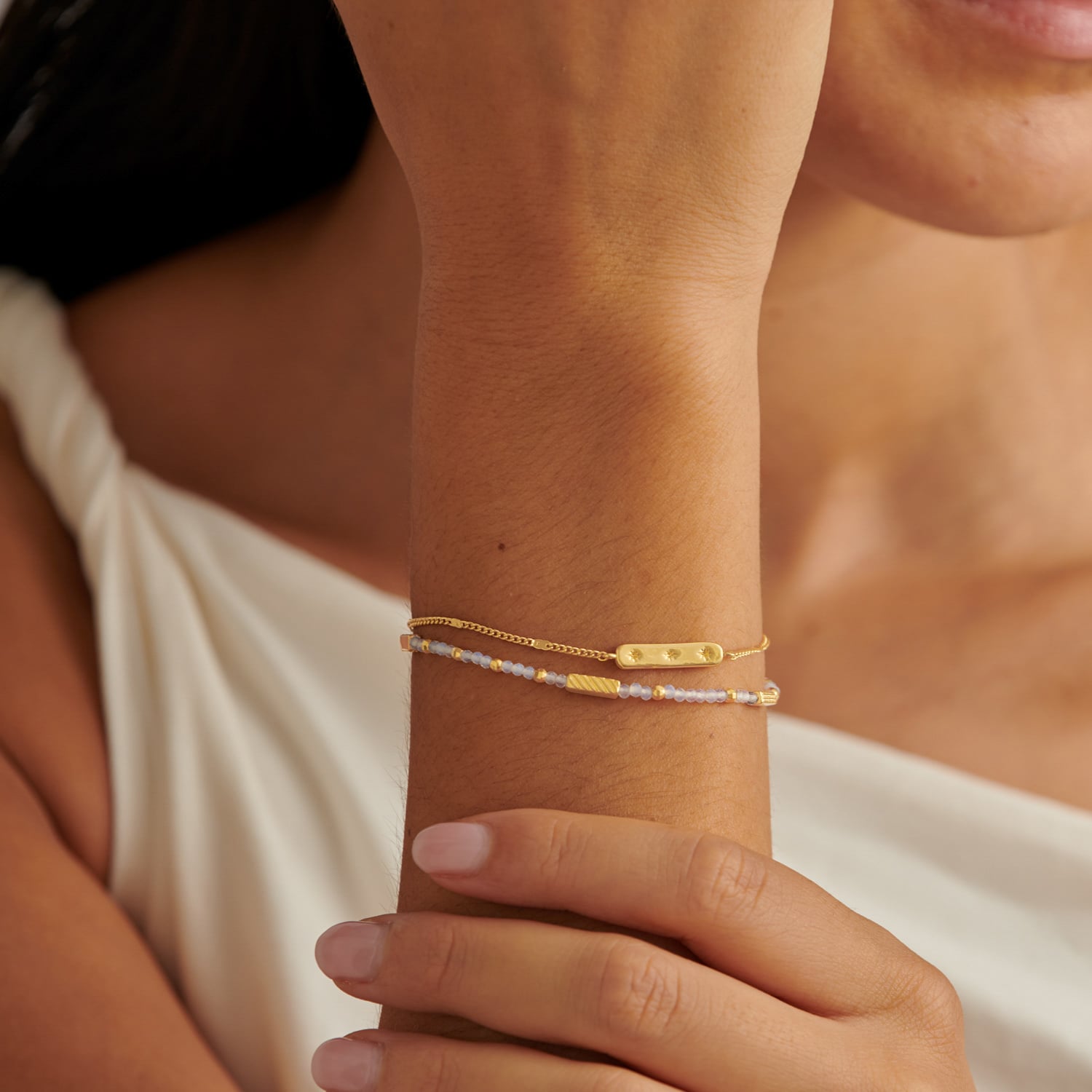 Two delicate gold bracelets worn on a wrist with small beads and a rectangular charm