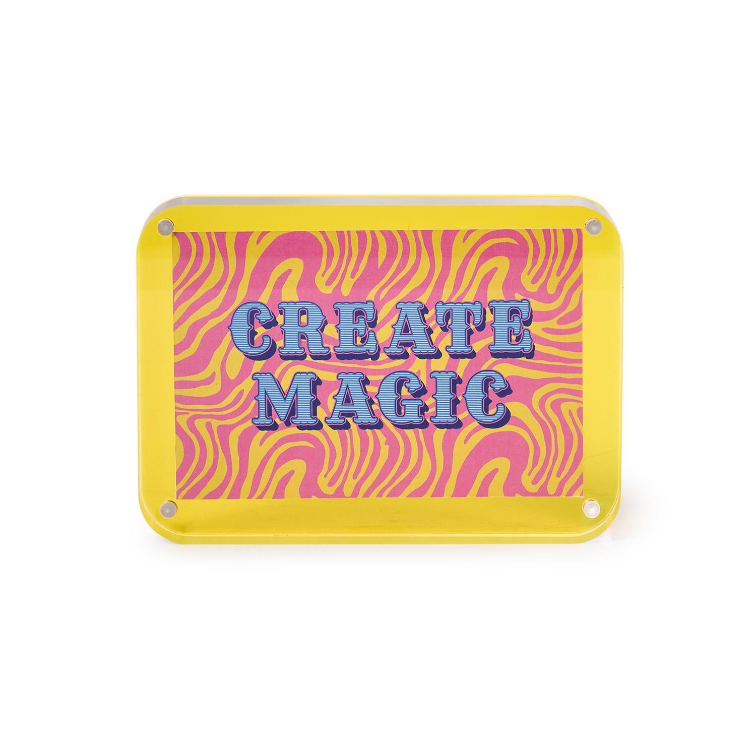A vibrant yellow acrylic sign featuring blue striped text CREATE MAGIC against a retro pink and yellow wavy background