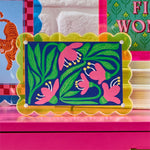 A vibrant modern floral art print with pink bell flowers and green leaves on a dark blue background, set in a unique scalloped yellow-orange iridescent acrylic frame, displayed on a bright pink surface alongside a light blue tiger print and a green Fi Won text print