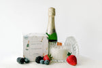 Bottle of champagne, a box labeled 'Champagne' next to a clear glass candle with a wooden wick and decorative items on a white surface.