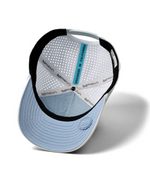 Product photograph by Melin of the interior of a white and light blue performance cap featuring a vertical teal strip reading DESIGNED IN CALIFORNIA, laser-perforated panels, and a light blue under-visor with a geometric pattern and a circular logo seal