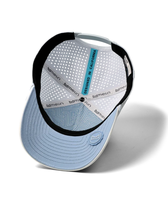 Product photograph by Melin of the interior of a white and light blue performance cap featuring a vertical teal strip reading DESIGNED IN CALIFORNIA, laser-perforated panels, and a light blue under-visor with a geometric pattern and a circular logo seal