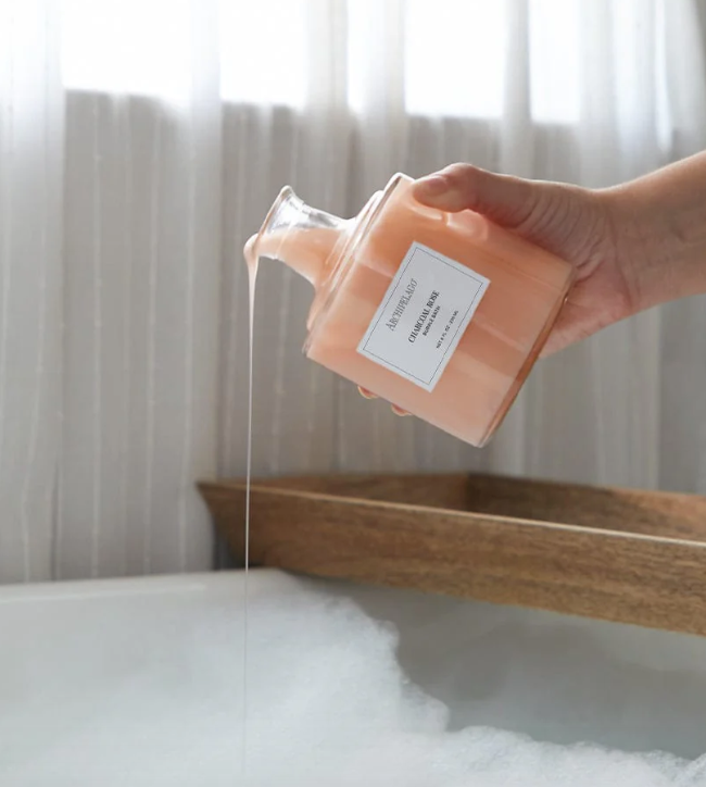Hand pouring pink bubble bath liquid from a glass bottle into a bathtub filled with foam