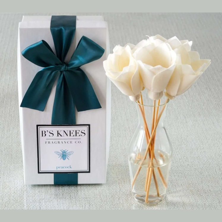 B'S Knees fragrance oil packaging with a teal ribbon and a vase of white flowers on a light background