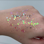 Photograph of holographic glitter swatch on a hand, featuring iridescent hexagonal and irregular flakes in pale pink, lime green, and gold, with the black text SWIFTIE overlaid