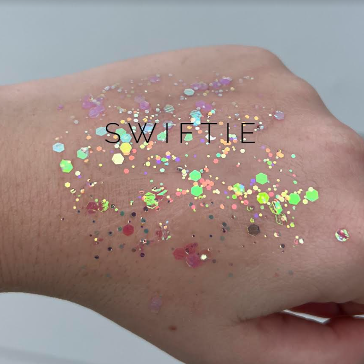 Photograph of holographic glitter swatch on a hand, featuring iridescent hexagonal and irregular flakes in pale pink, lime green, and gold, with the black text SWIFTIE overlaid