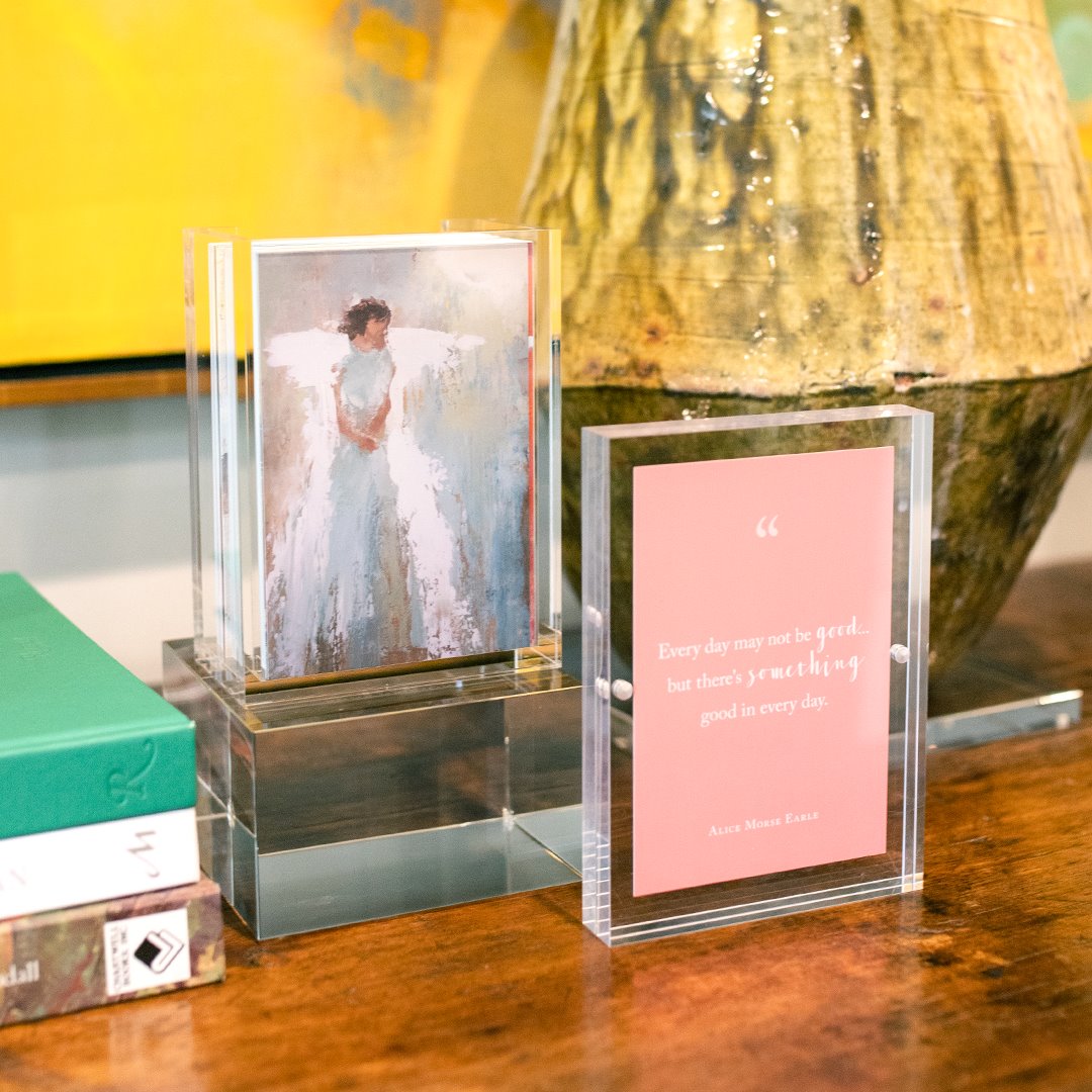 Acrylic frames displaying an art print of a figure and a pink card with an inspirational quote on a wooden surface