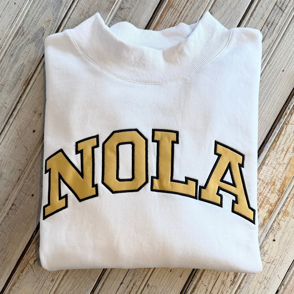 White sweatshirt with gold and black NOLA lettering on a wooden surface