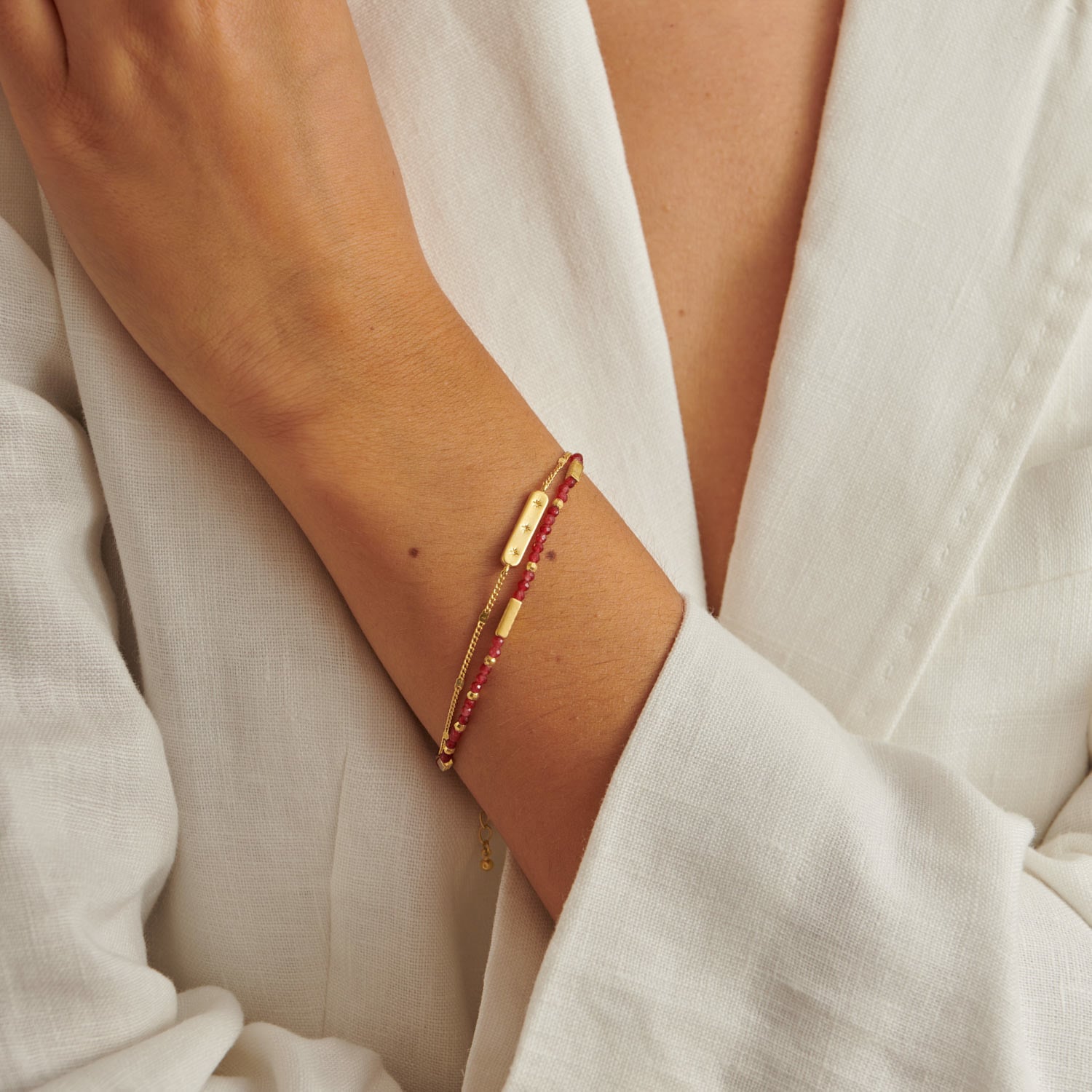 Gold chain bracelet with a small rectangular charm and a second strand of red beaded gemstones on a wrist