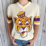 Person wearing a LSU-themed sports jersey with a tiger logo on a wooden background
