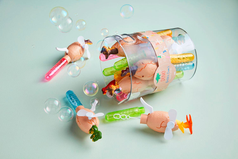 Photograph of dinosaur egg-shaped bubble fans featuring mini white propellers and colorful solution tubes in pink, blue, green, and orange, spilling from a 'DINO Bubble Fans' package onto a light blue surface among iridescent bubbles