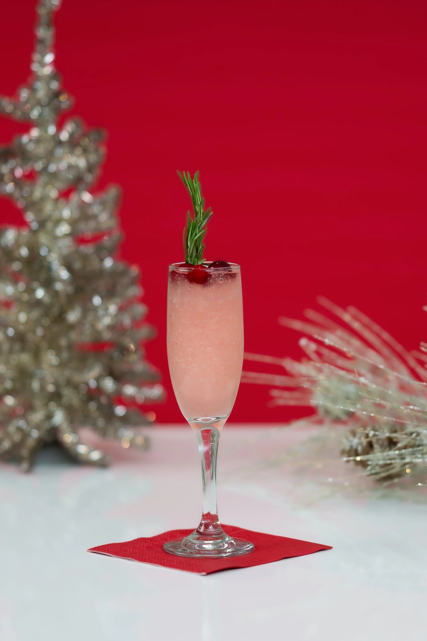 Pink cocktail in a champagne flute garnished with rosemary and cranberries on a red napkin