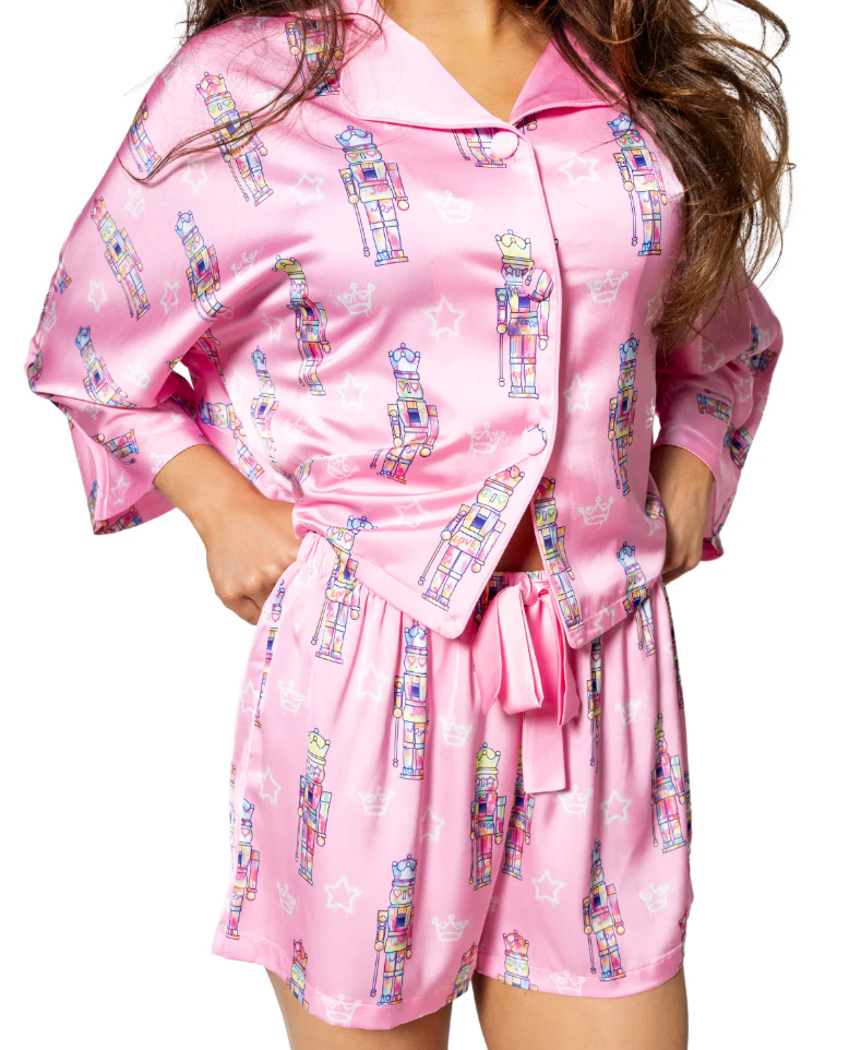 Photograph of a person wearing a bright pink satin pajama set featuring a repeating pattern of colorful stylized nutcracker soldiers, white stars, and outlines of crowns, with a button-up top and shorts tied with a bow