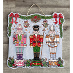 Door hanger with three nutcrackers in pink, red and white under holly garland and red bows