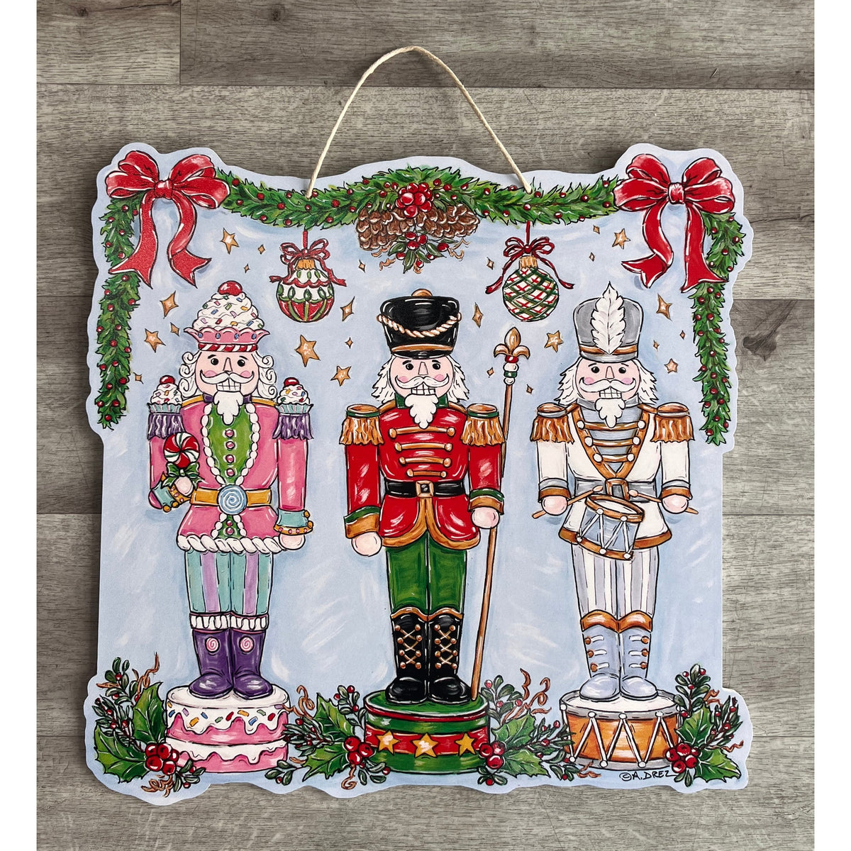 Door hanger with three nutcrackers in pink, red and white under holly garland and red bows