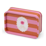 Product photography by Paddywax of an Orange Blossom Soy Wax Candle in a rectangular tin featuring vibrant horizontal pink and orange stripes and a scalloped oval label with a pink sun face motif indicating 6.5 oz 184g