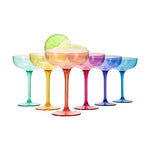 A row of six colorful acrylic coupe glasses, including teal, yellow, orange, magenta, deep blue, and light blue, with the orange glass holding a light green drink and a lime wedge garnish, all on a crisp white background