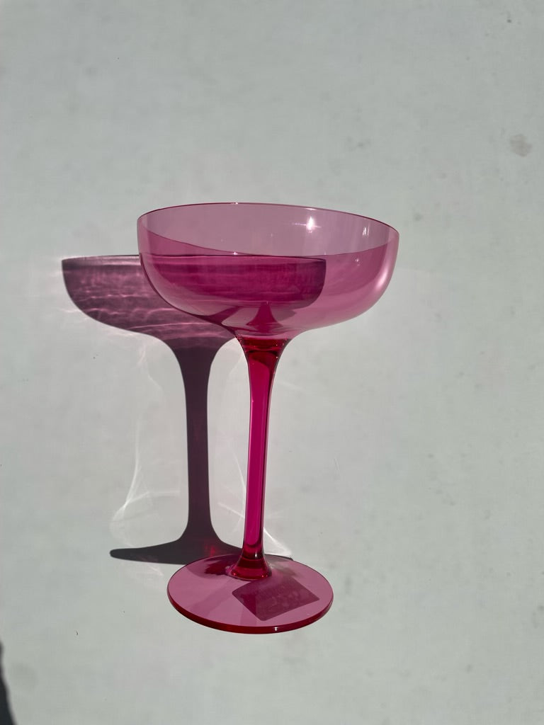 Photograph of a vibrant pink coupe glass standing on a bright white surface, casting a long dark shadow with a distinct rippled light reflection