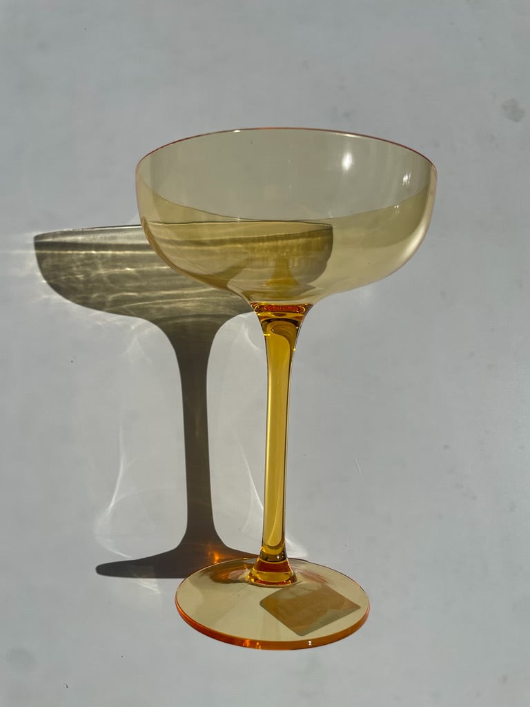 Photograph of a single amber gold coupe glass on a light background casting a striped shadow with a sticker visible on its base