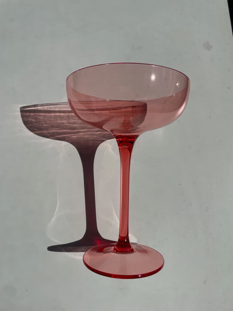 Photograph of a vibrant pink coupe glass standing on a light surface casting a sharp, elongated shadow with subtle light reflections