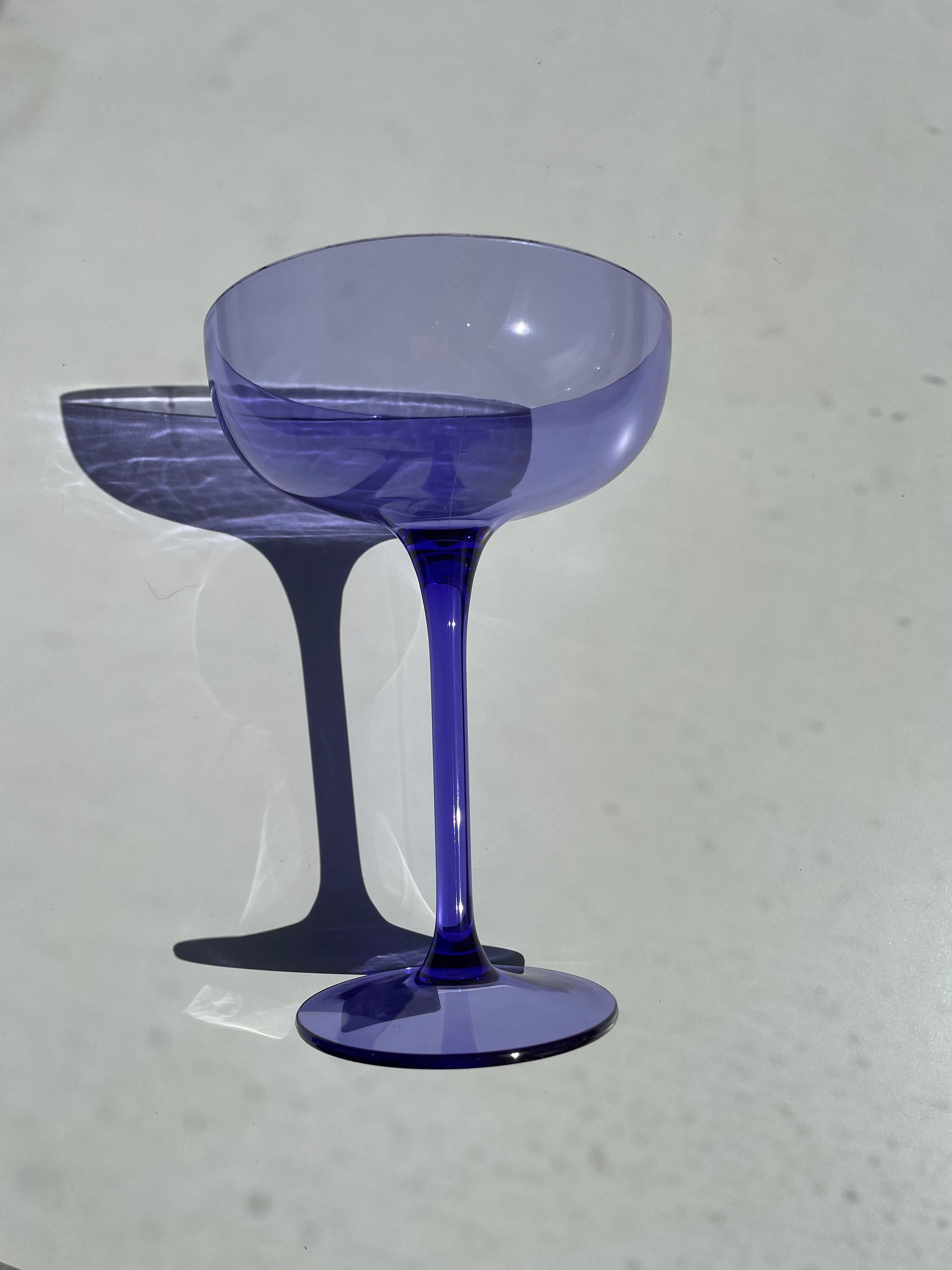 Photograph of a single deep purple coupe glass on a stark white surface casting a sharp shadow with light refracting through its amethyst-colored bowl and stem