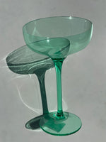 A translucent emerald green coupe glass on a plain grey surface casting a long dramatic shadow and intricate light refractions