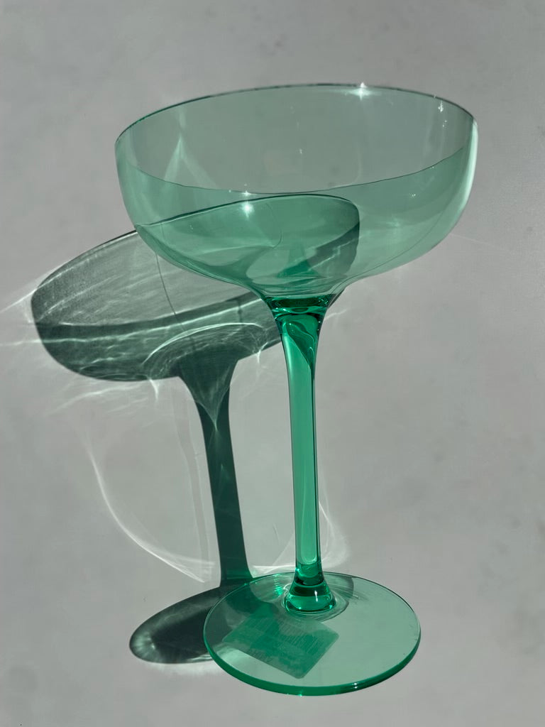 A translucent emerald green coupe glass on a plain grey surface casting a long dramatic shadow and intricate light refractions