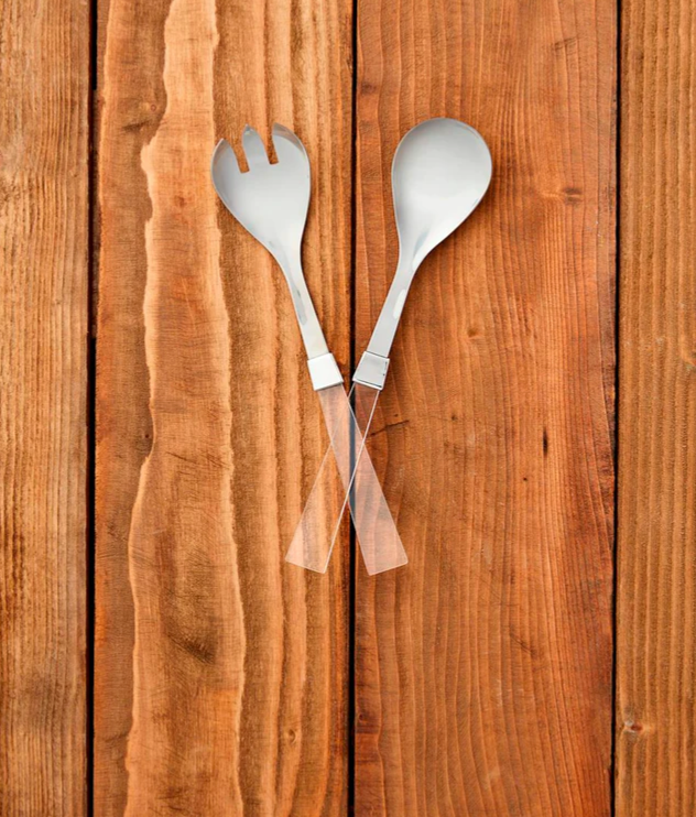 Stainless steel salad servers with clear acrylic handles crossed on a wooden surface